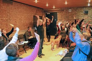 Bridget stands facing seated participants - all reach one arm straight up above their heads, hand held with fingers joined to create a beak