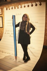 Photo shows a young woman with a giant pencil. She stands on what appears to be a sheet from a notebook, enlarged 25 times. Notes on the page refer to 'scale'.